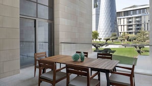 Un área de estar exterior moderna con una mesa y sillas de madera, ventanas amplias y vista a un edificio con una torre geométrica distintiva.