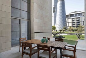 Un área de estar exterior moderna con una mesa y sillas de madera, ventanas amplias y vista a un edificio con una torre geométrica distintiva.