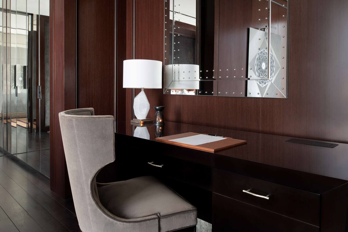 Una habitación de hotel moderna con un espejo grande, un escritorio con lámpara, una silla acolchada y una tocador de madera.