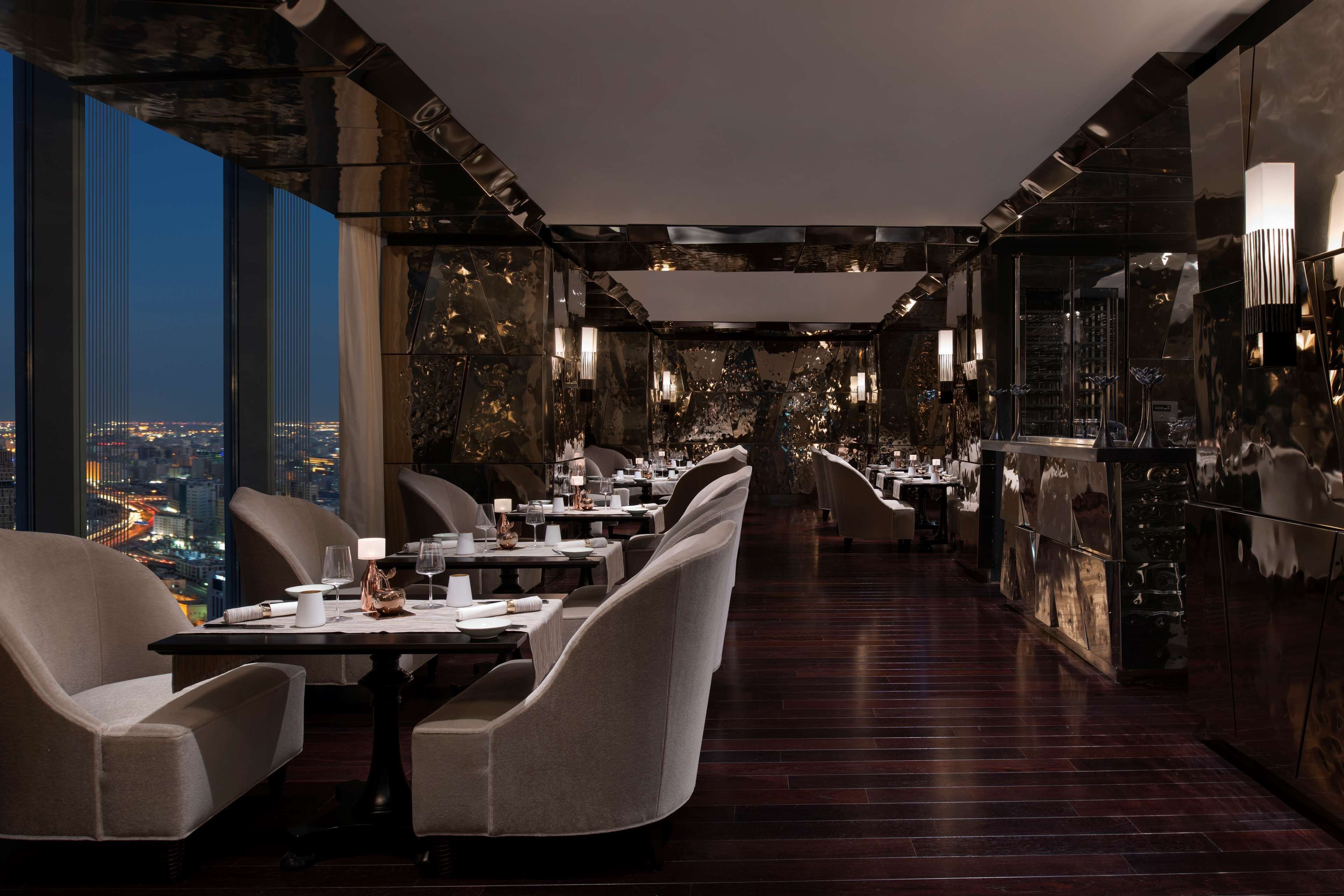 A modern restaurant with a long dining area, high ceilings, and a city view.