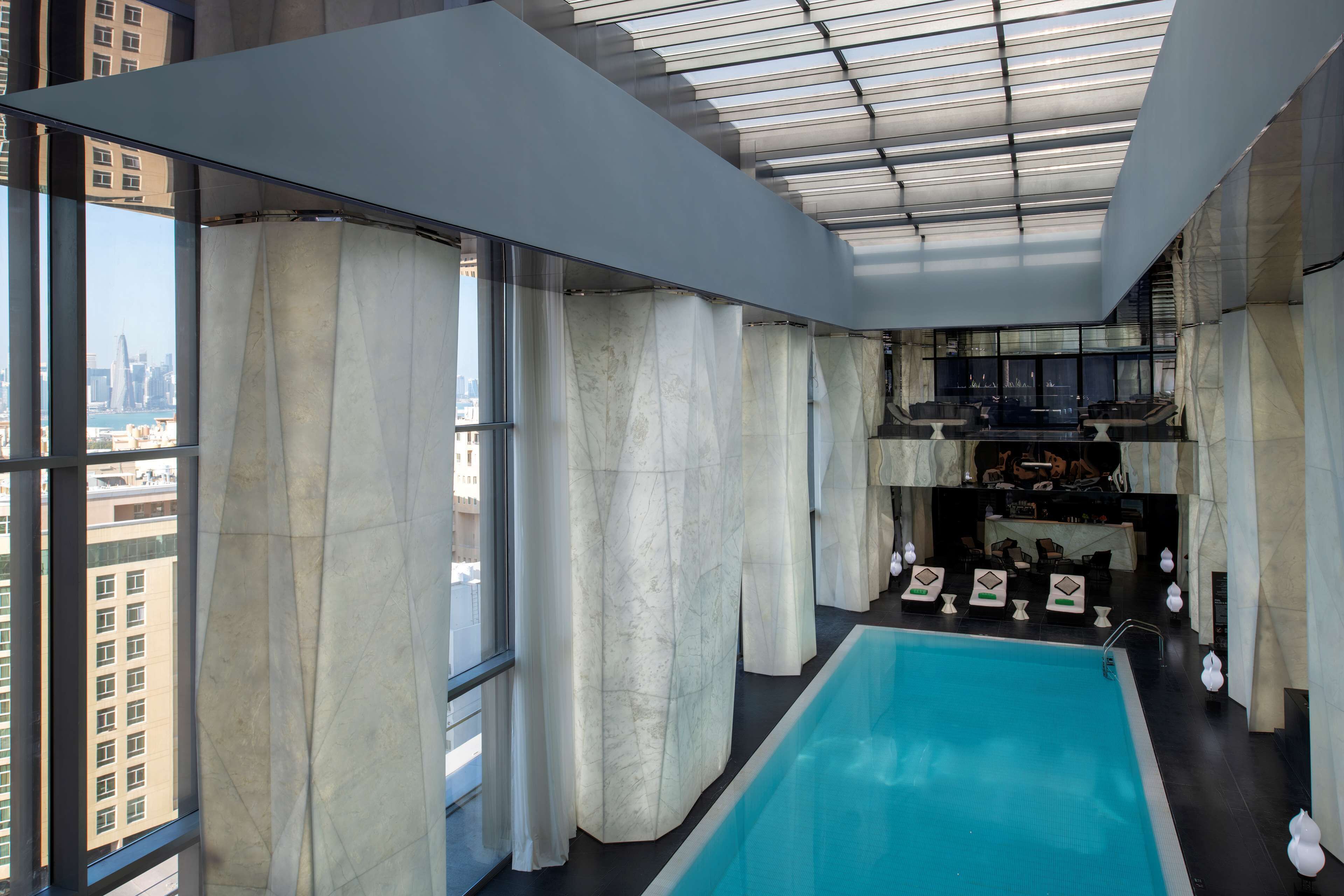 A modern indoor swimming pool with a city view, large windows, and a bar area.