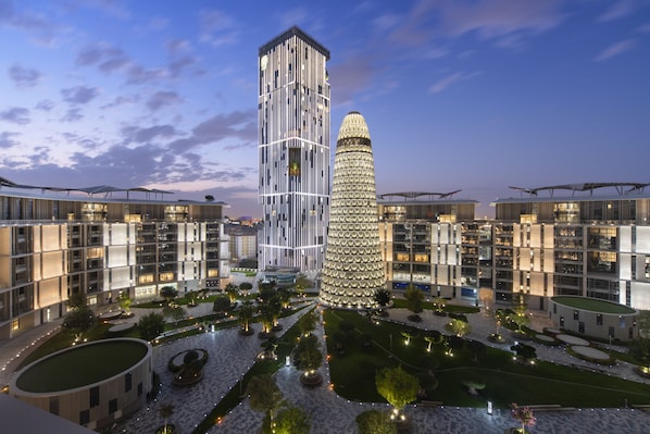 Un complejo hotelero moderno con una torre central y edificios circundantes, que cuenta con un patio con jardines ajardinados y senderos.