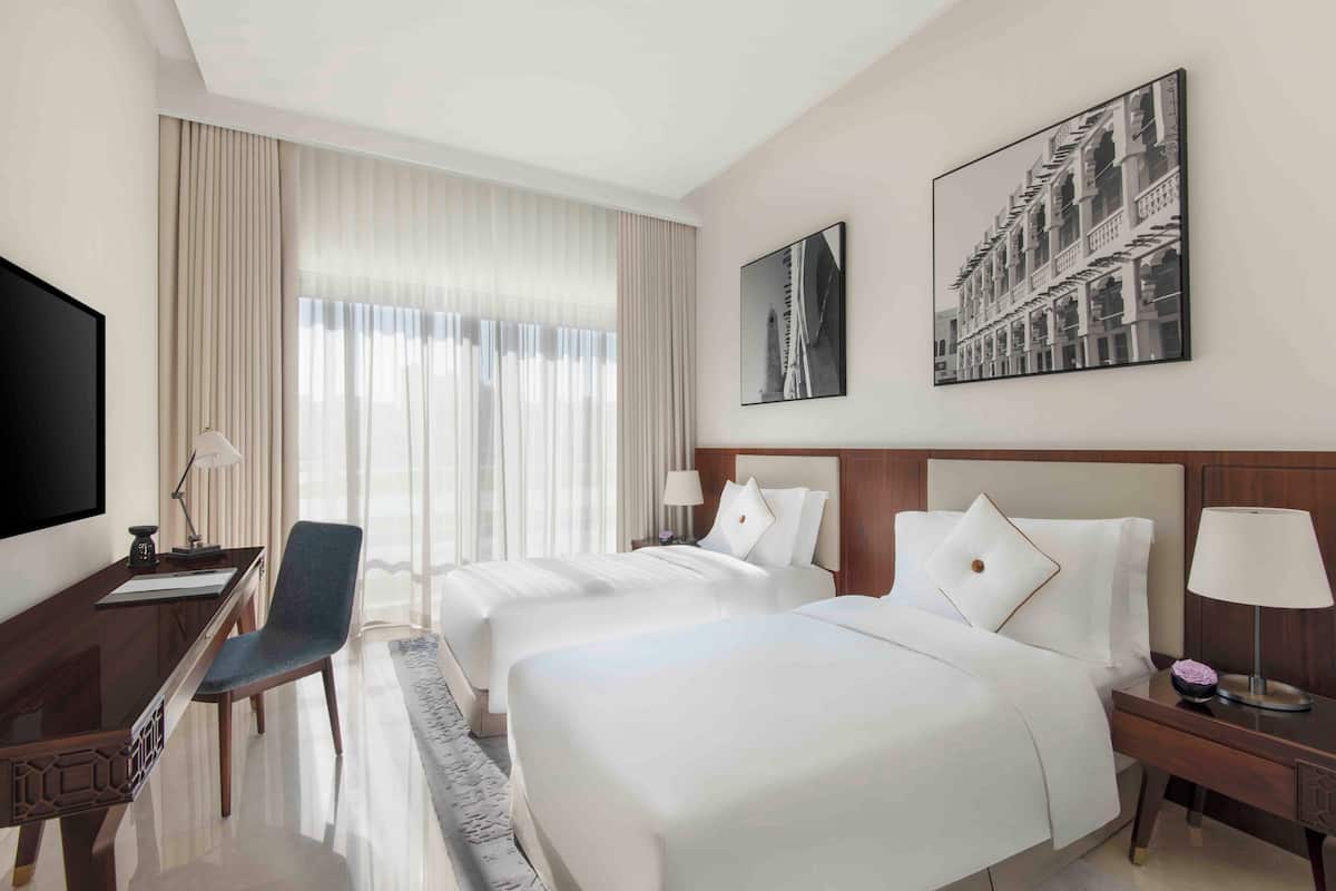 Ein Hotelzimmer mit zwei Betten, einem Schreibtisch, einem Stuhl, einem Fernseher und Bildern an den Wänden.