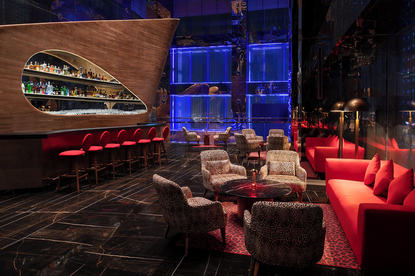 A bar area with a curved wooden counter, red bar stools, leopard print chairs, and a red sofa.