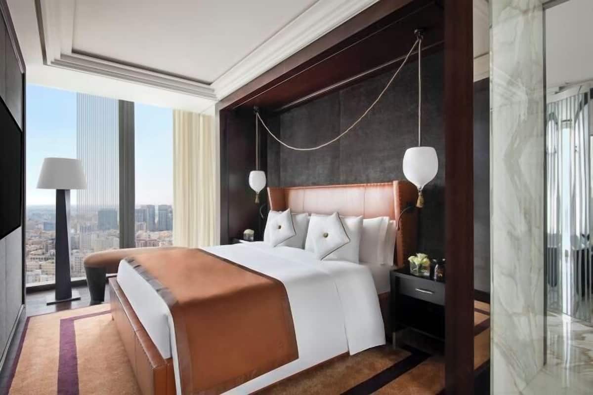 Ein Hotelzimmer mit einem großen Bett, einem Nachttisch, einer Lampe und Blick auf die Stadt.