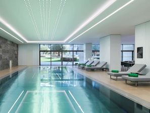 Ein modernes Hallenbad mit großen Glaswänden, einer Steinwand und Liegestühlen mit grünen Kissen.