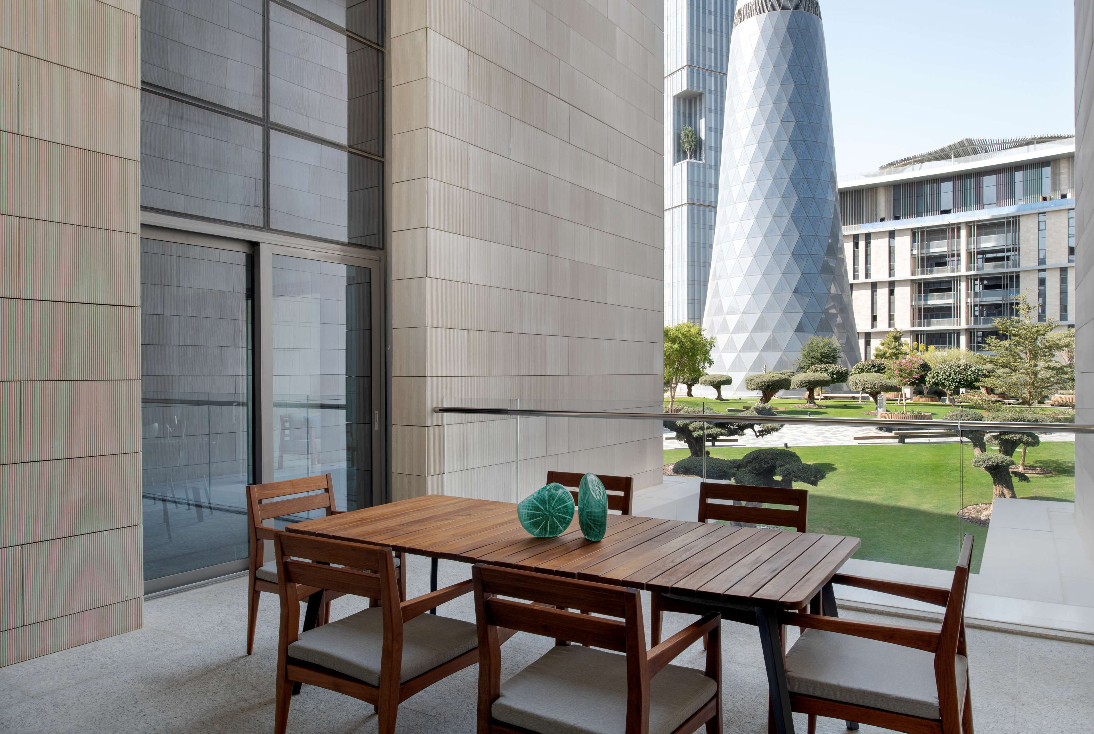A modern outdoor seating area with a wooden table and chairs, a glass wall, and a view of a building with a unique geometric design.