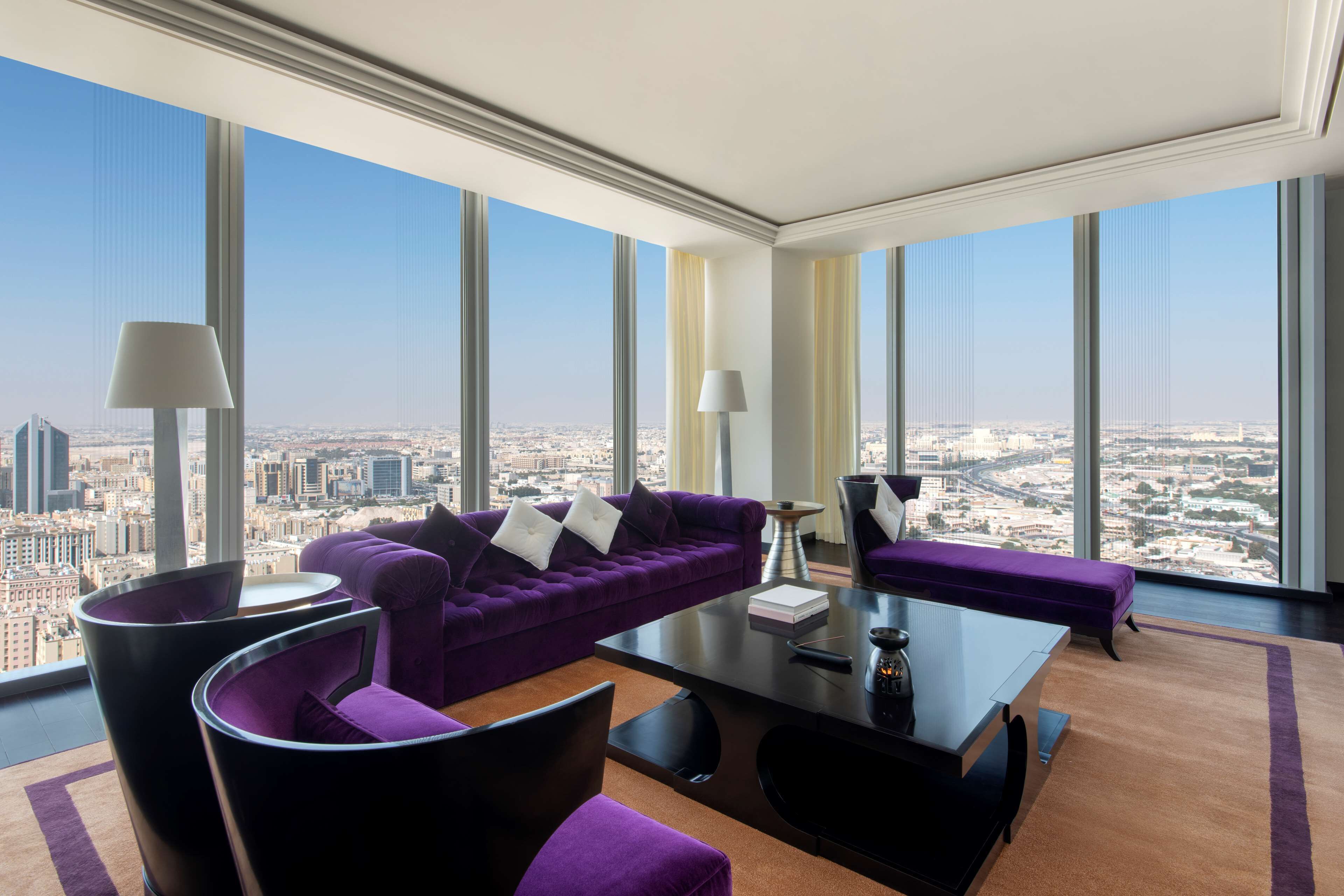 A high-rise hotel room with a city view, a purple sofa, a black coffee table, and a modern lamp.