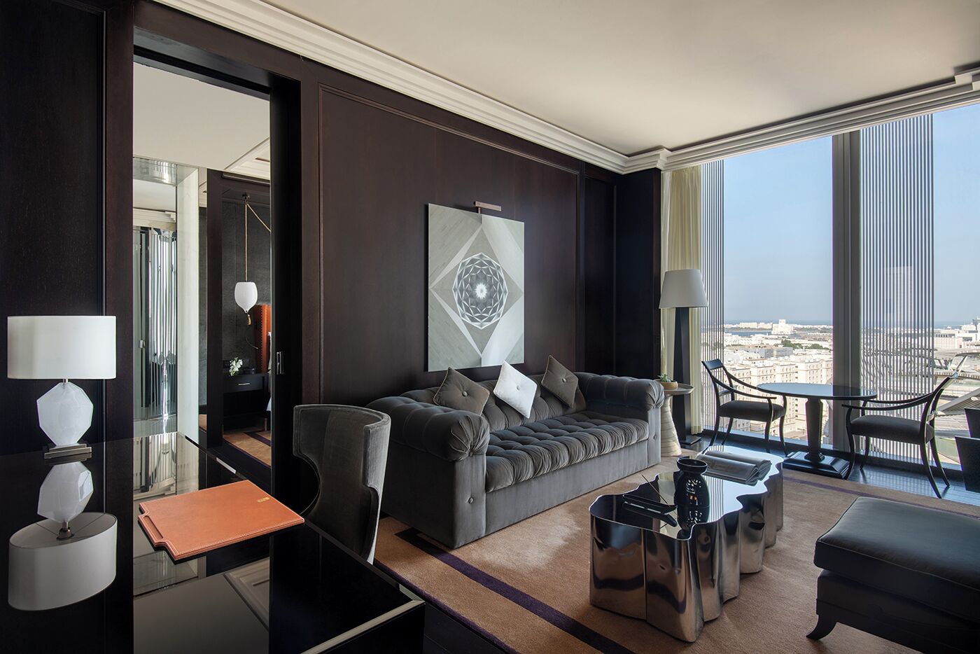 Habitación de hotel moderna con pared de madera oscura, un ventanal con vista a la ciudad, un sofá gris con cojines, una mesita redonda y una mesa de centro de vidrio.