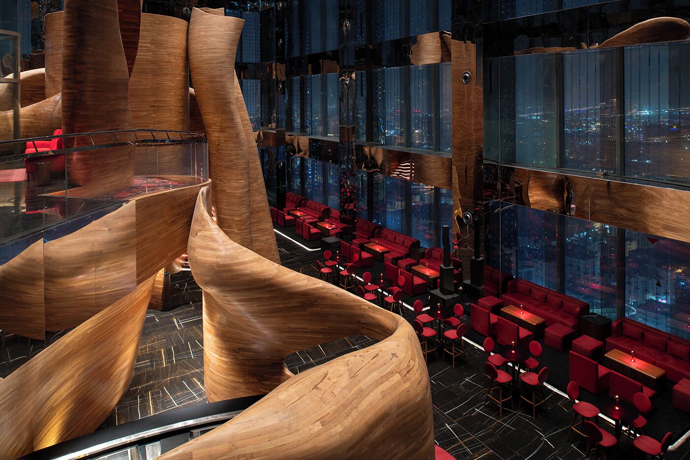 A modern interior with a spiral wooden structure, red seating, and large windows offering city views.