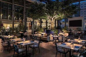 An outdoor dining area with tables and chairs, surrounded by potted plants and a large tree. - Banyan Tree Doha at La Cigale Mushaireb (Doha)