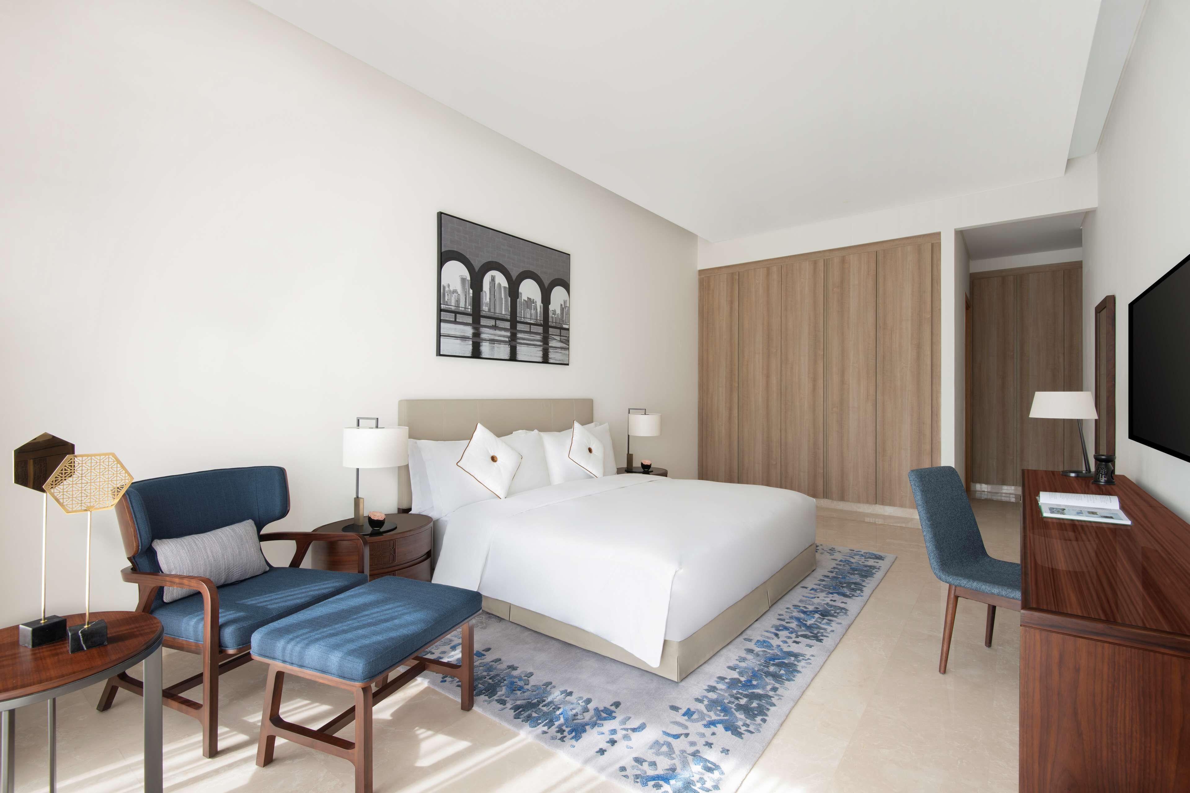 A hotel room with a bed, a desk, a chair, a bedside table, a lamp, a TV, and a painting on the wall.