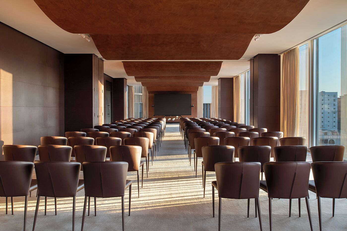 A conference room with rows of chairs facing a black screen, large windows, and a modern ceiling design.