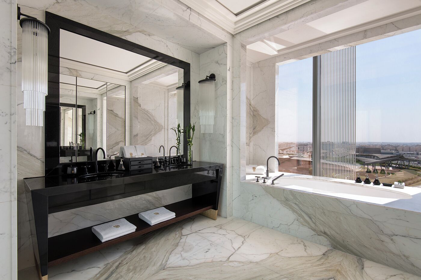 A modern bathroom with a large mirror, black vanity, marble flooring, and a view of the city through a large window.