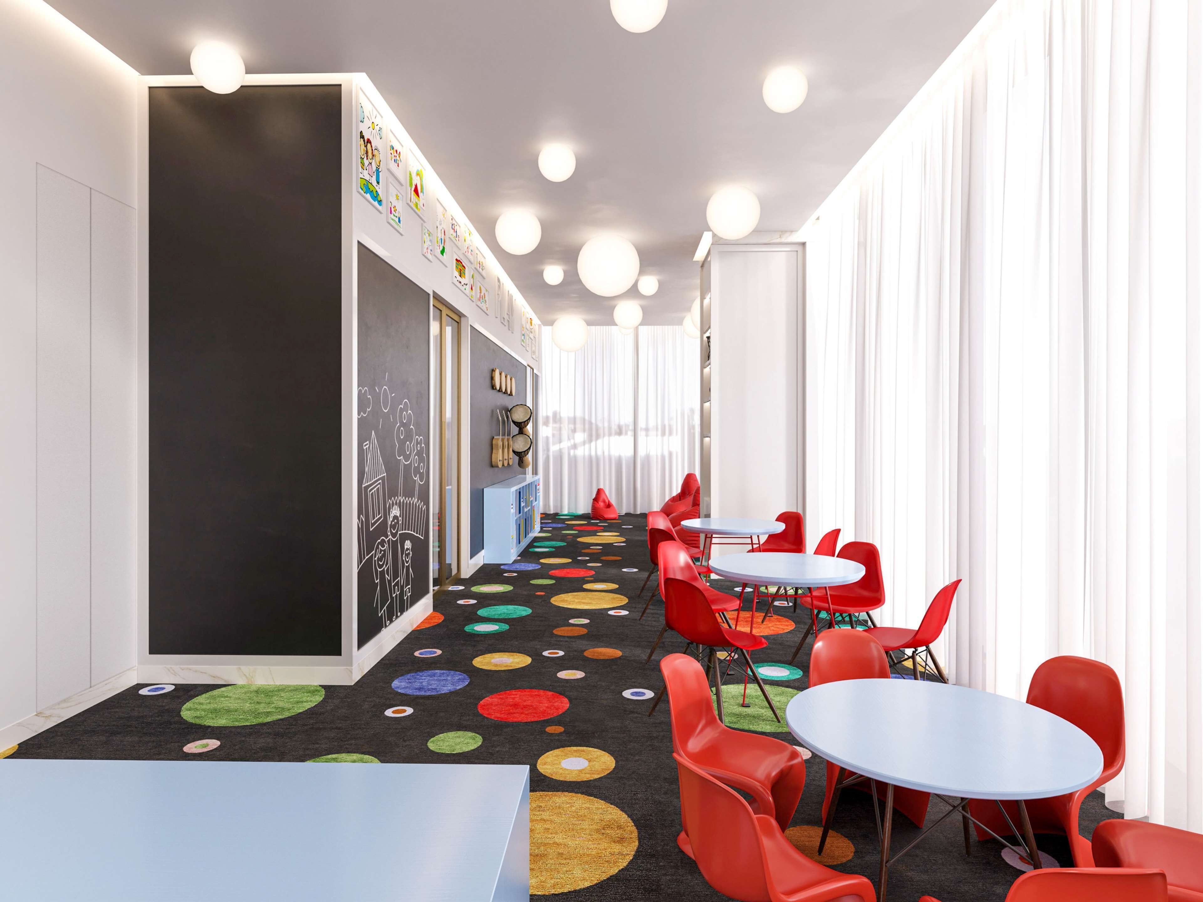 Un salón de juegos infantiles con un suelo circular de colores, sillas rojas y una pared de pizarra llena de dibujos.