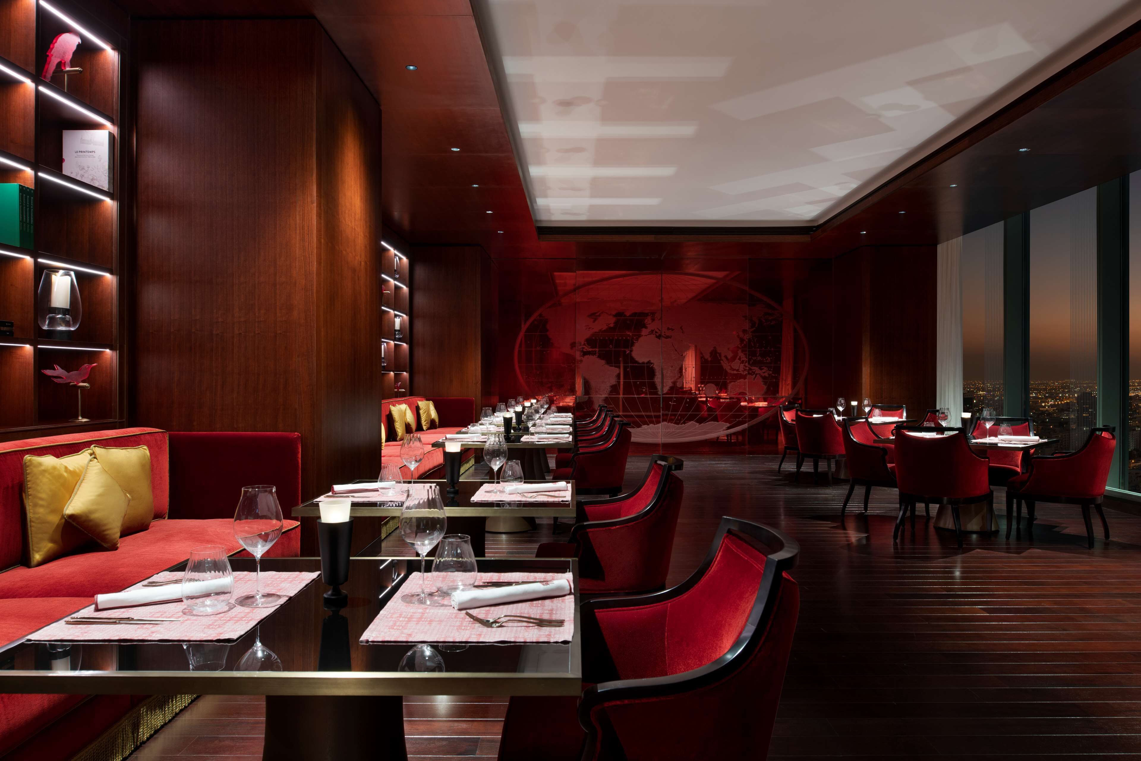 Un restaurant doté de nombreuses tables, de chaises à l’assise rembourrée de tissu rouge et offrant une vue sur le paysage urbain grâce à de grandes fenêtres.