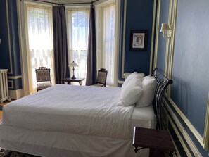 A large bed with white linens, a nightstand, a lamp, and a chair in a room with blue walls and a window with curtains.