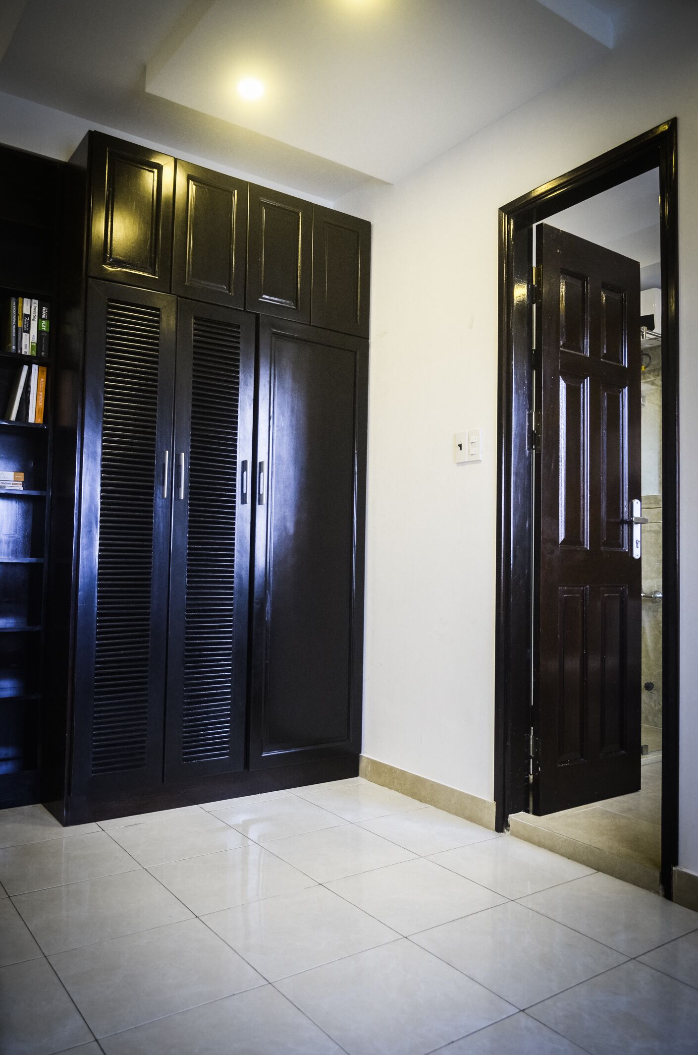 A room with a dark wooden wardrobe, a white tiled floor, and a partially open door leading to another room.