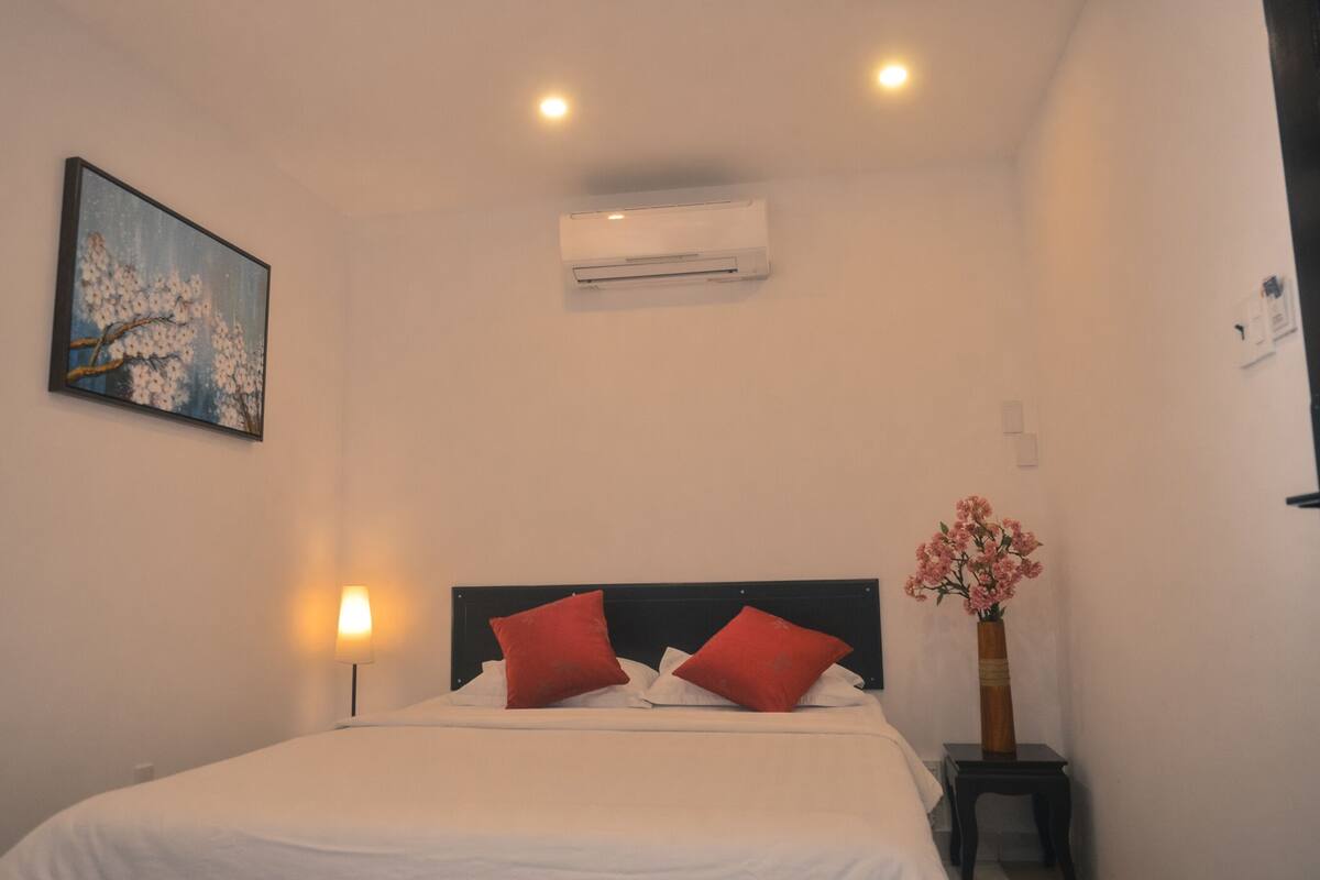 Una cama bien hecha con ropa de cama blanca y dos almohadas rojas, un equipo de aire acondicionado montado en la pared, una lámpara de noche y un jarrón con flores rosas sobre una mesita de noche.