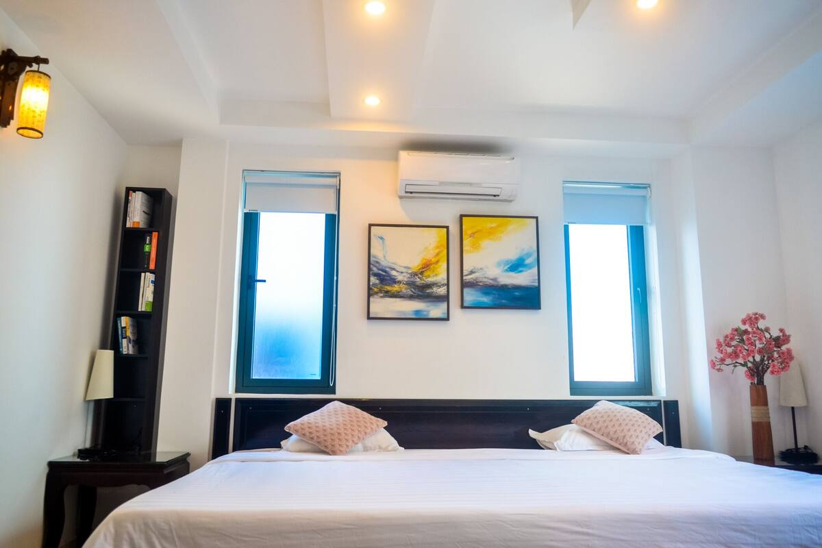 Habitación de hotel con una cama grande, dos ventanas, equipo de aire acondicionado, una estantería y un florero con flores.