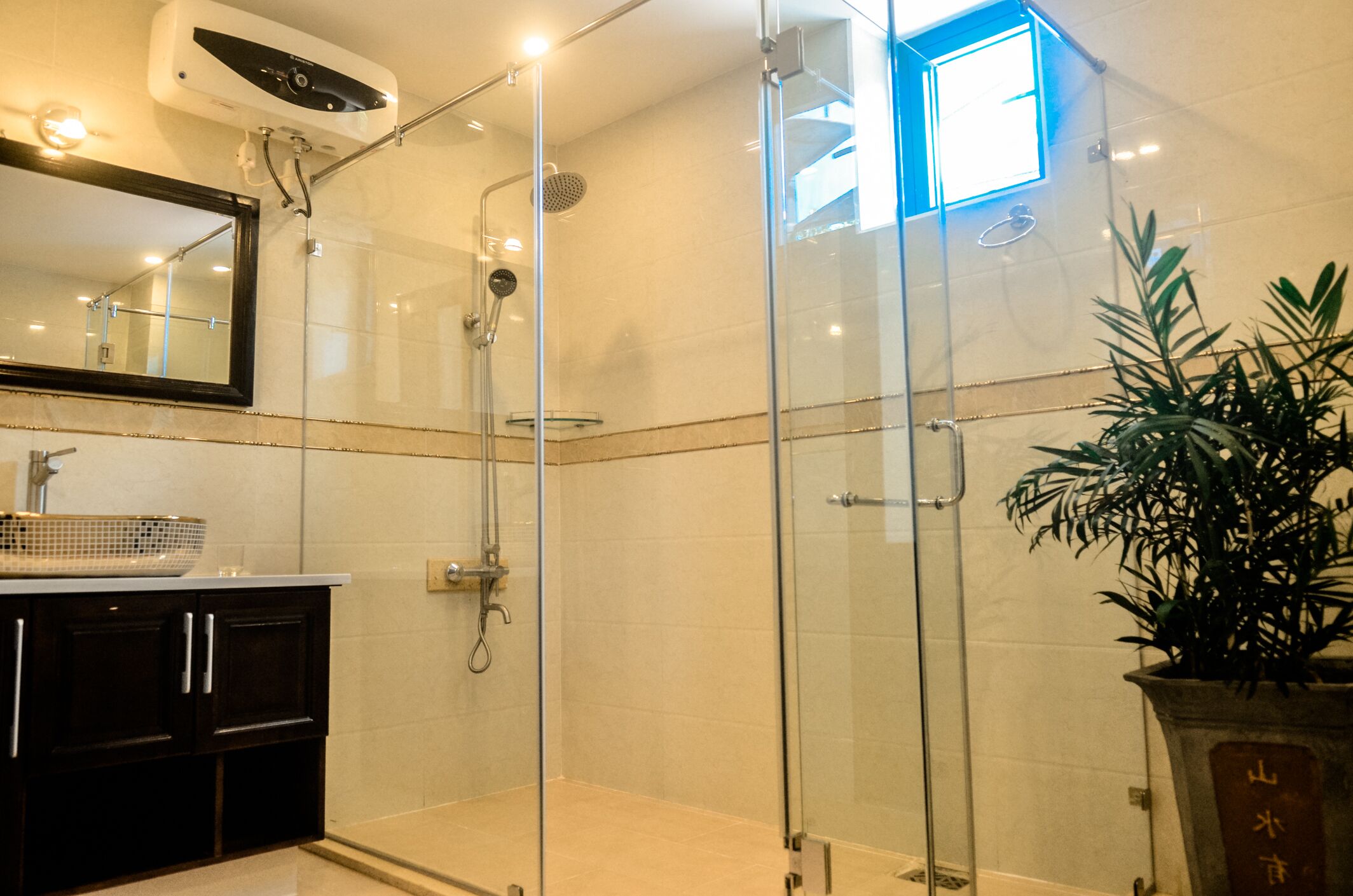 A modern bathroom with a glass-enclosed shower, a large mirror, and a potted plant.