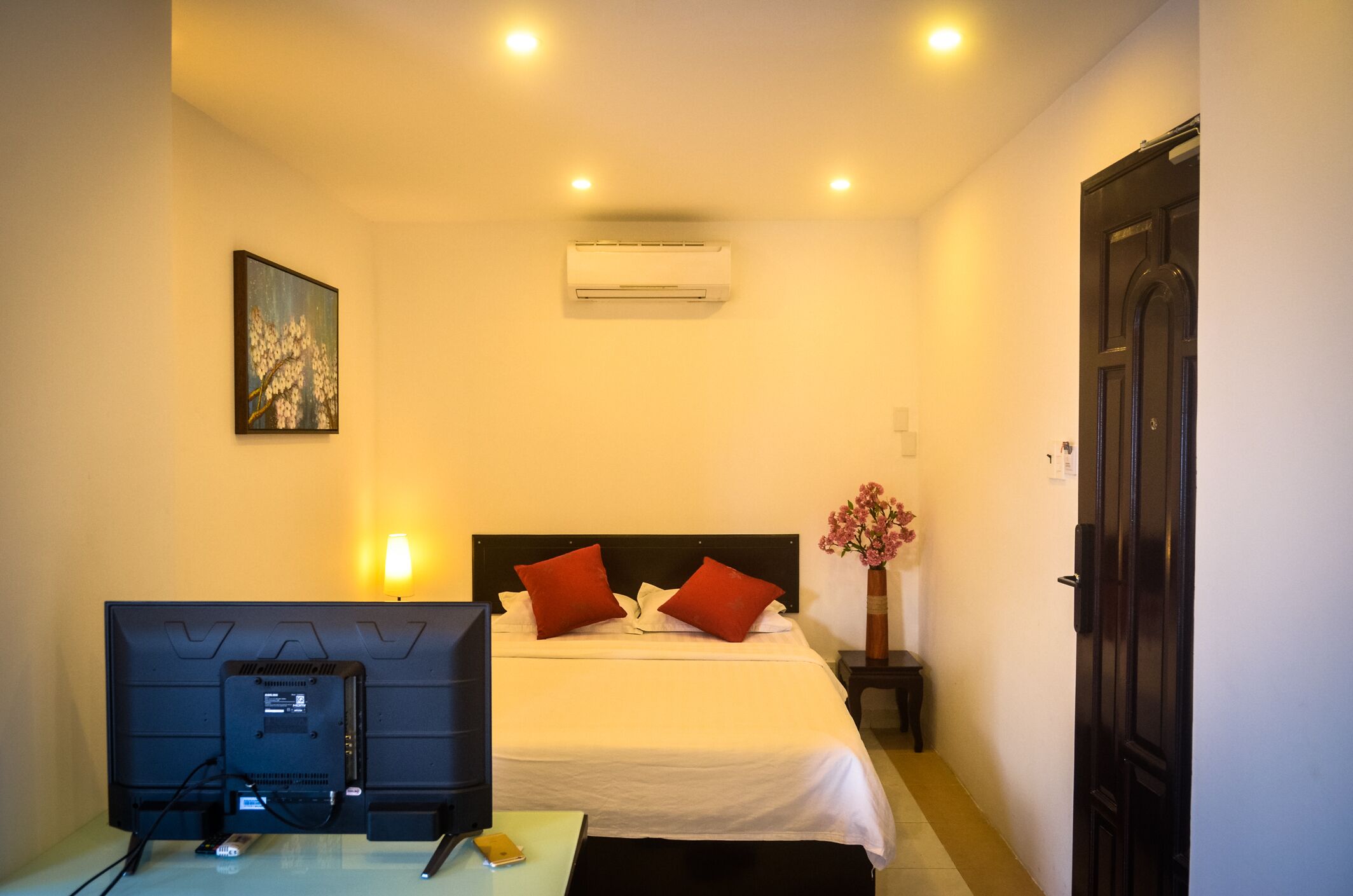 A hotel room with a bed, a flat-screen TV, an air conditioner, and a vase with flowers.