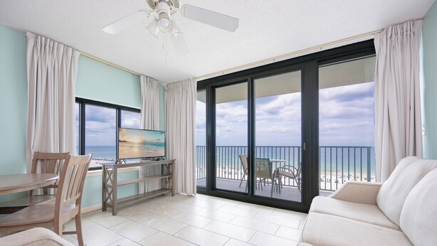 A room with a dining area, a sofa, a TV, and a balcony with a view of the beach.