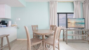 A room with a round wooden table, four chairs, a TV on a stand, a window with curtains, and a kitchenette with a coffee maker.