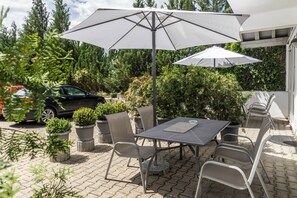 An outdoor patio with a table and chairs, a large umbrella, potted plants, and a car in the background.