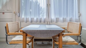 Ein Zimmer mit Tisch und zwei Stühlen, einem Fenster mit transparenten Vorhängen, einem Heizkörper und einer Tür mit Rauchverbot-Hinweis.