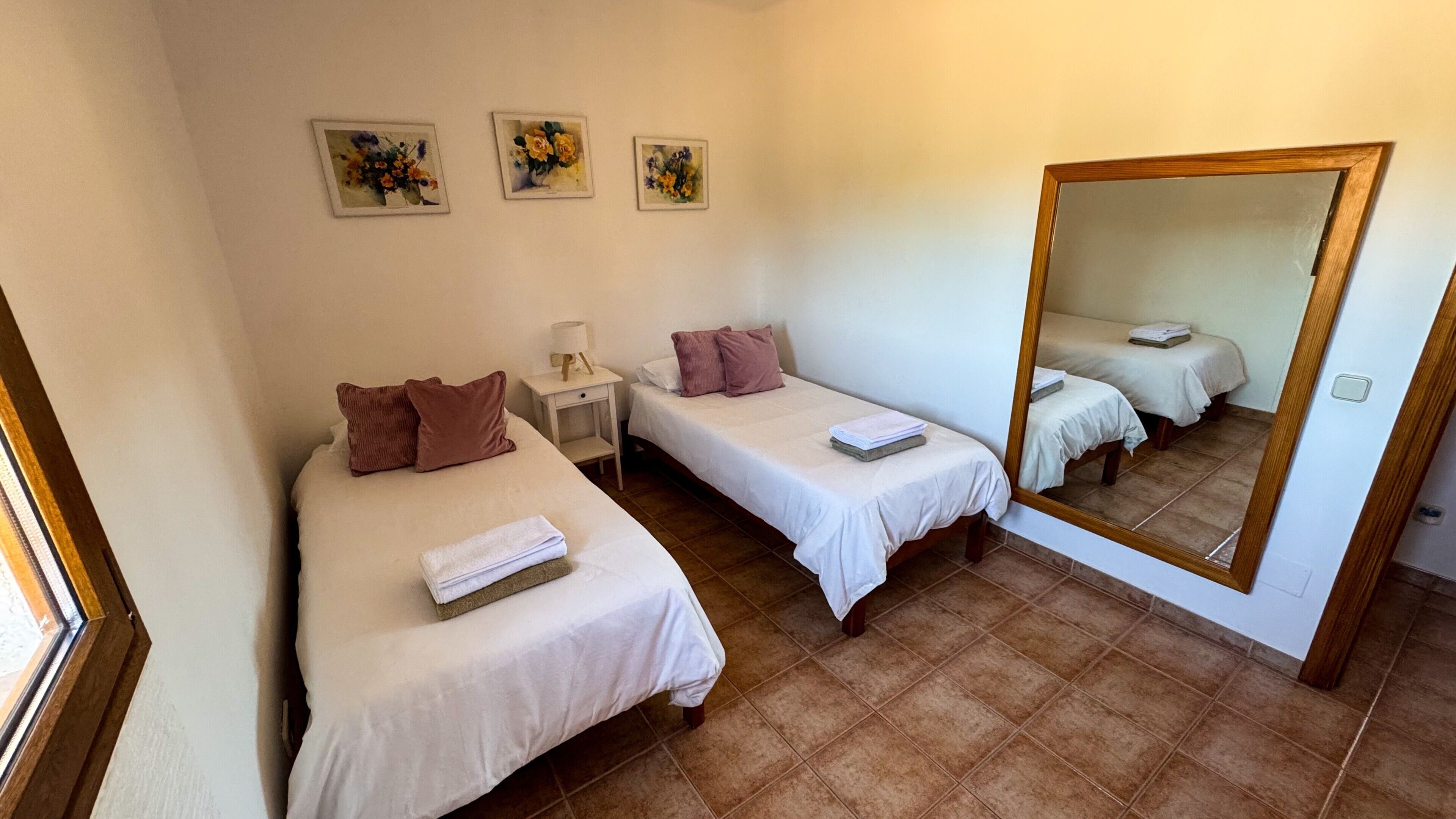 A room with two single beds, a nightstand with a lamp, a mirror, and three floral paintings on the wall.