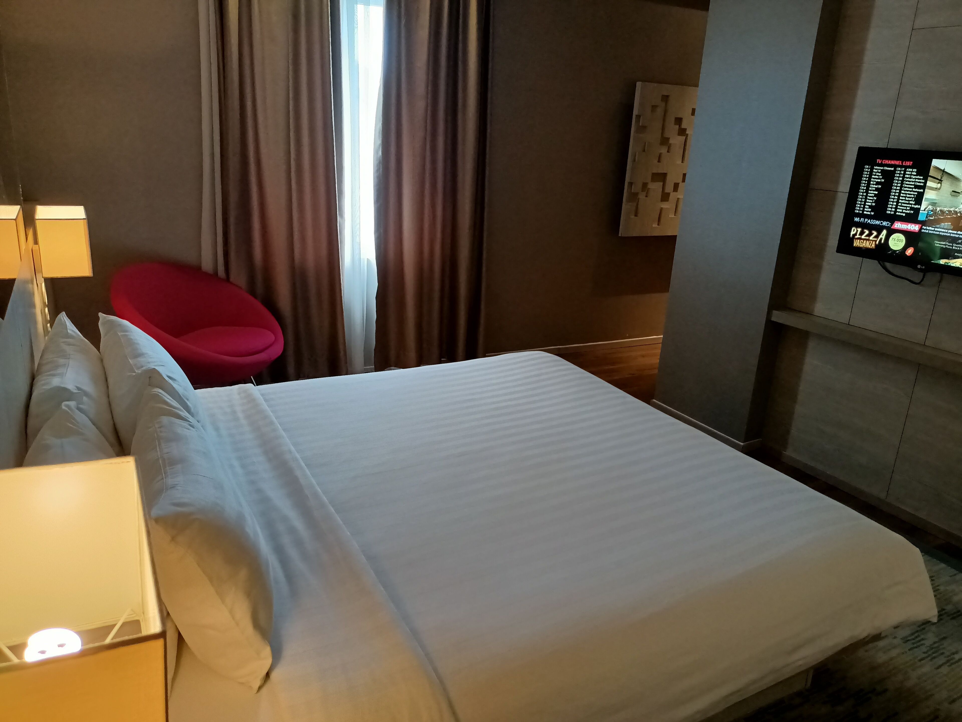 Habitación de hotel con cama, una silla roja, televisor montado en la pared y una ventana con cortinas.