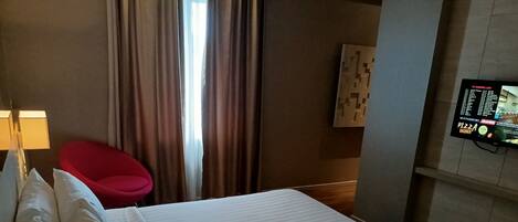 Habitación de hotel con cama, una silla roja, televisor montado en la pared y una ventana con cortinas.