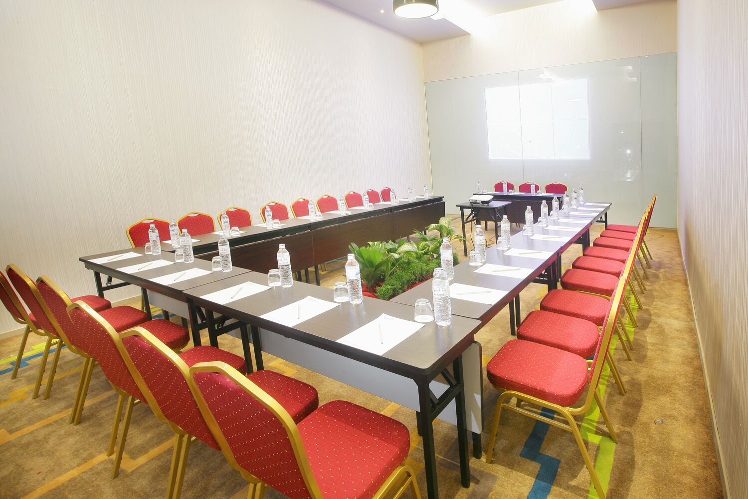 Una sala conferenze con un lungo tavolo, sedie rosse, bottiglie d'acqua e uno schermo per proiezioni.