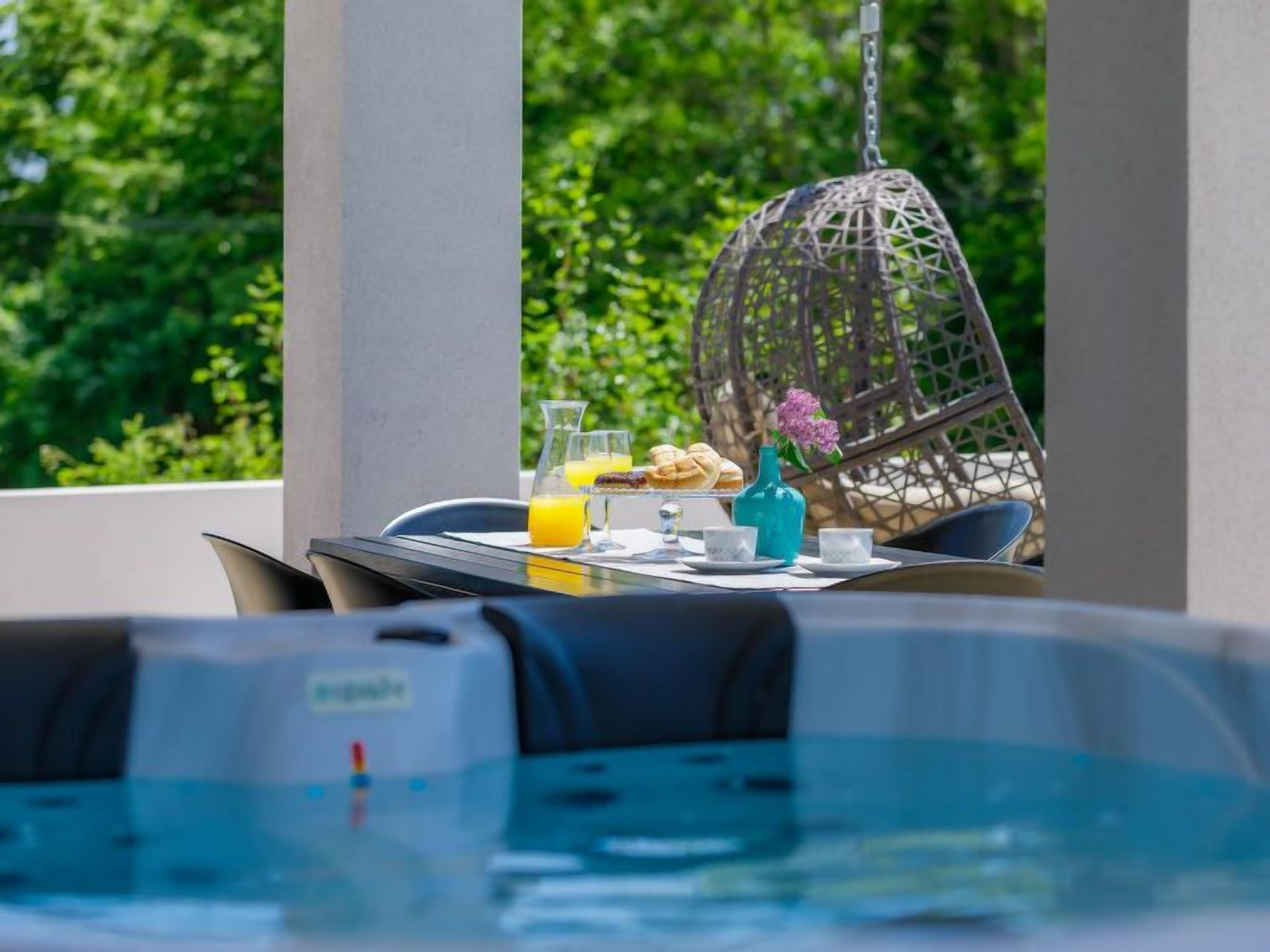 A poolside dining area with a table set for breakfast, including croissants and orange juice, and a hanging chair in the background.