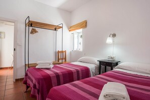 Una habitación con dos camas, un tendedero de madera, una silla, una mesita de noche y una lámpara de pared.
