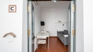 A room with a white desk, a chair, a grey sofa, a TV mounted on the wall, and a small shelf.