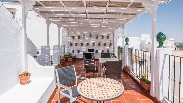 Una terraza con una pérgola blanca, plantas en macetas, mobiliario exterior y vistas de la ciudad.