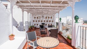 Een terras met een witte pergola, planten in potten, tuinmeubilair en uitzicht op de stad.
