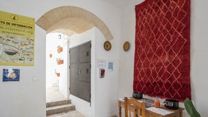 A hallway with a tiled floor, a wooden chair, a table with a red patterned cloth, a door with a metal gate, and a poster on the wall. - Holiday Apartment "Casa La Costanilla D" with Roof Terrace and Air Conditioning (Vejer de la Frontera)