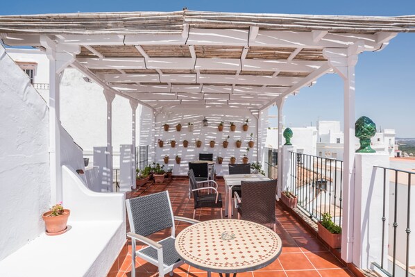 Una terraza con una pérgola blanca, plantas en macetas, mobiliario exterior y vistas de la ciudad.