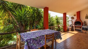 Une terrasse extérieure avec une table recouverte d’une nappe violette, entourée de chaises, et un four en briques rouges.