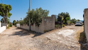 Une allée de gravier mène à une entrée fermée par un portail, avec un mur en béton et des plantes en pot.