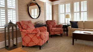 Un salon avec un plafond en bois, un miroir rond, deux fauteuils à motifs, un canapé beige et une table basse.