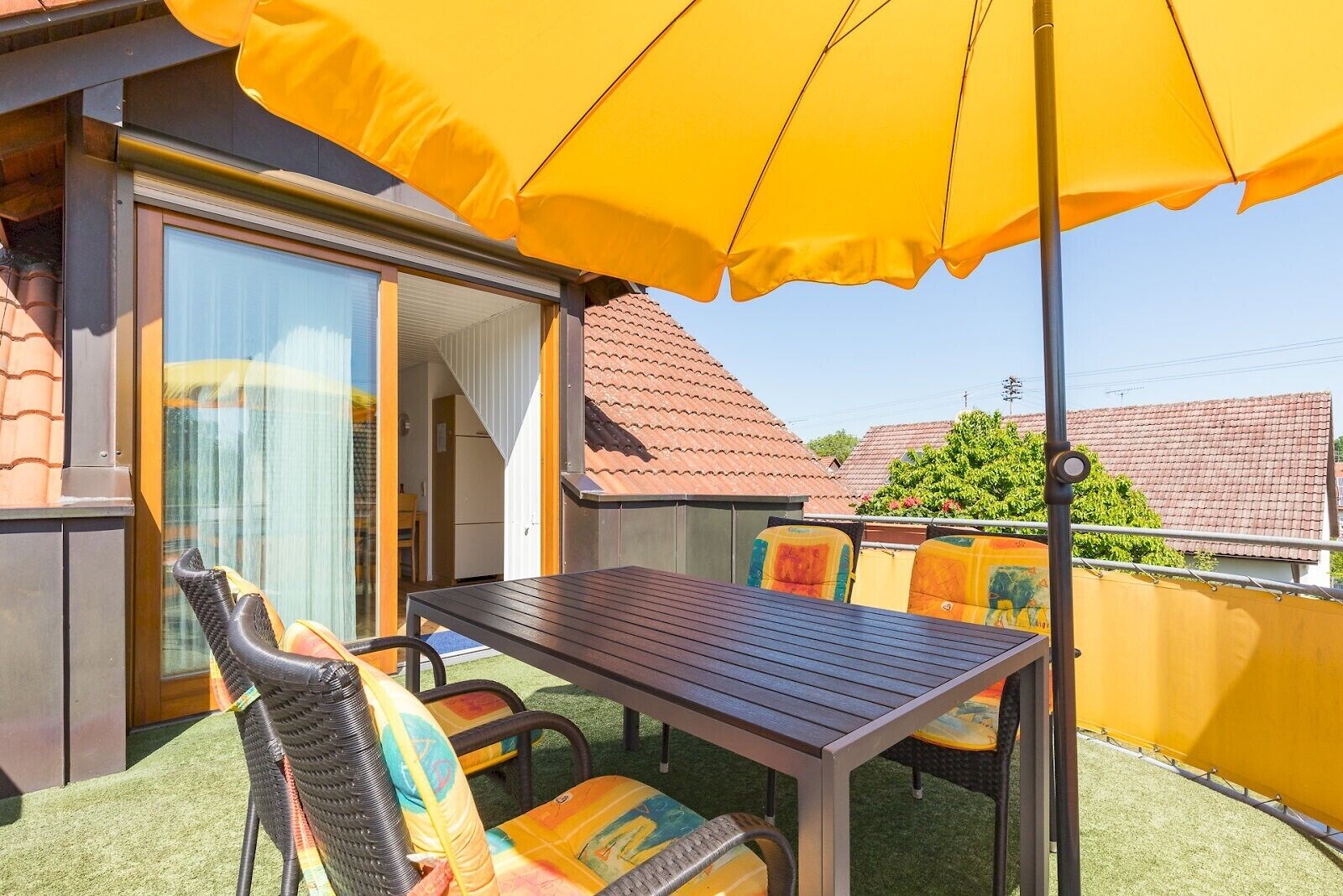 Eine Terrasse im Freien mit Tisch und Stühlen, einem gelben Sonnenschirm und einer Glasschiebetür, die zu einem Zimmer führt.