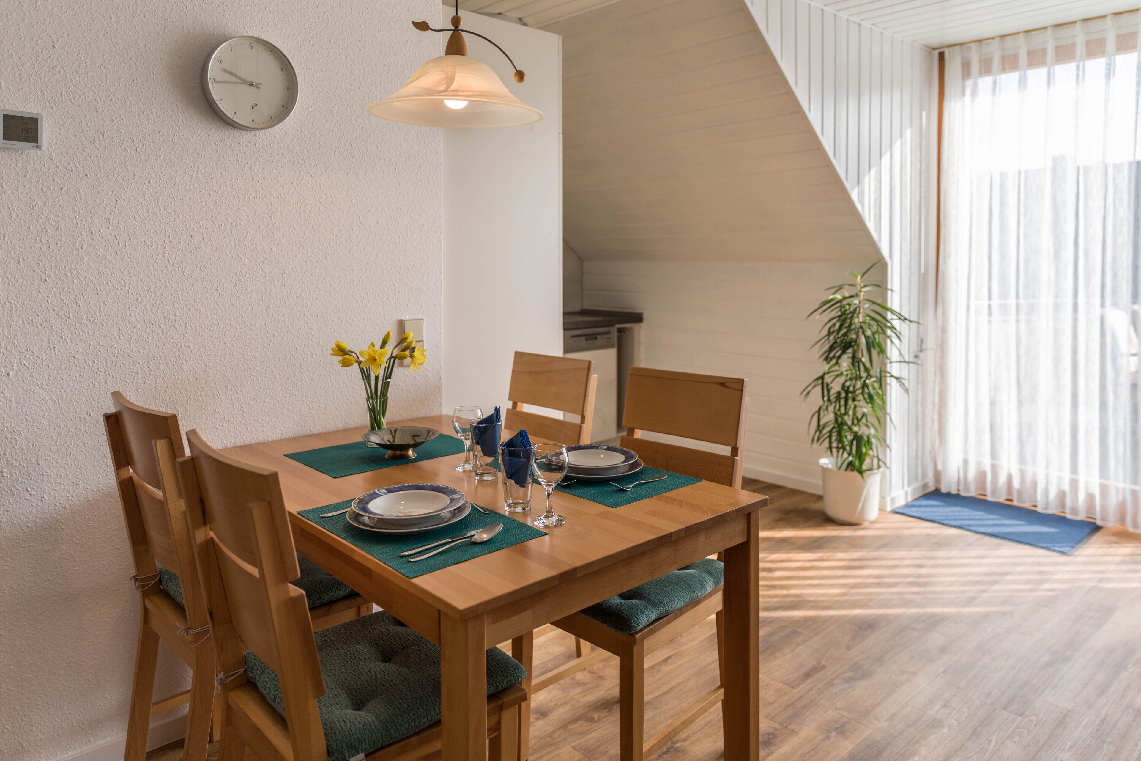 Ein Essbereich mit einem Holztisch, gedeckt für vier Personen, Stühle mit grünen Kissen, einer Uhr an der Wand und einem großen Fenster mit transparenten Vorhängen.