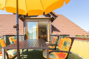 A balcony with a table and chairs, a yellow umbrella, and a view of a building with a tiled roof.