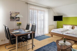 A hotel room with a dining table, chairs, a bed with a green headboard, a TV, and a painting on the wall.