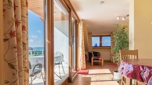 Una habitación con puerta de vidrio corrediza que da a un balcón, una mesa de madera, una silla, una planta en maceta y una ventana con vistas.