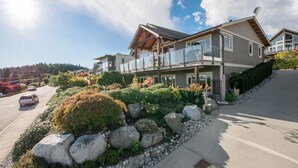 A modern two-story house with a balcony, a driveway, and a well-maintained garden with rocks and shrubs.